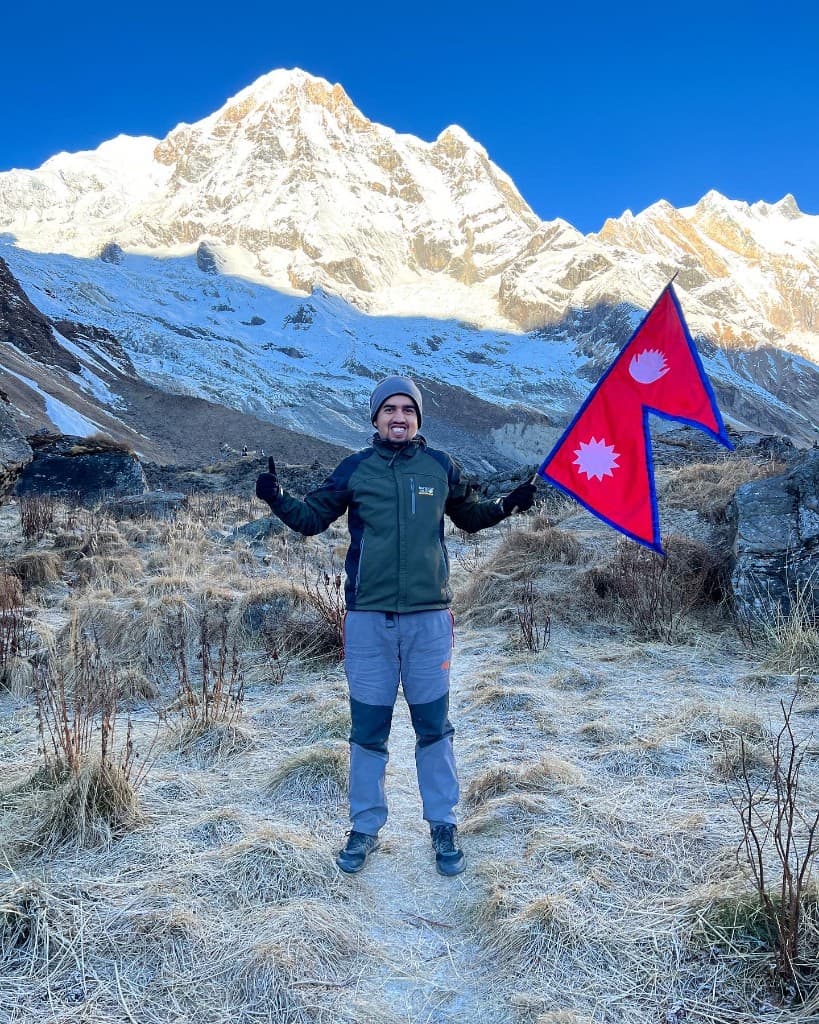 Pratap Sharma holding a Nepali flag at Annapurna Base Camp with frost on the grass