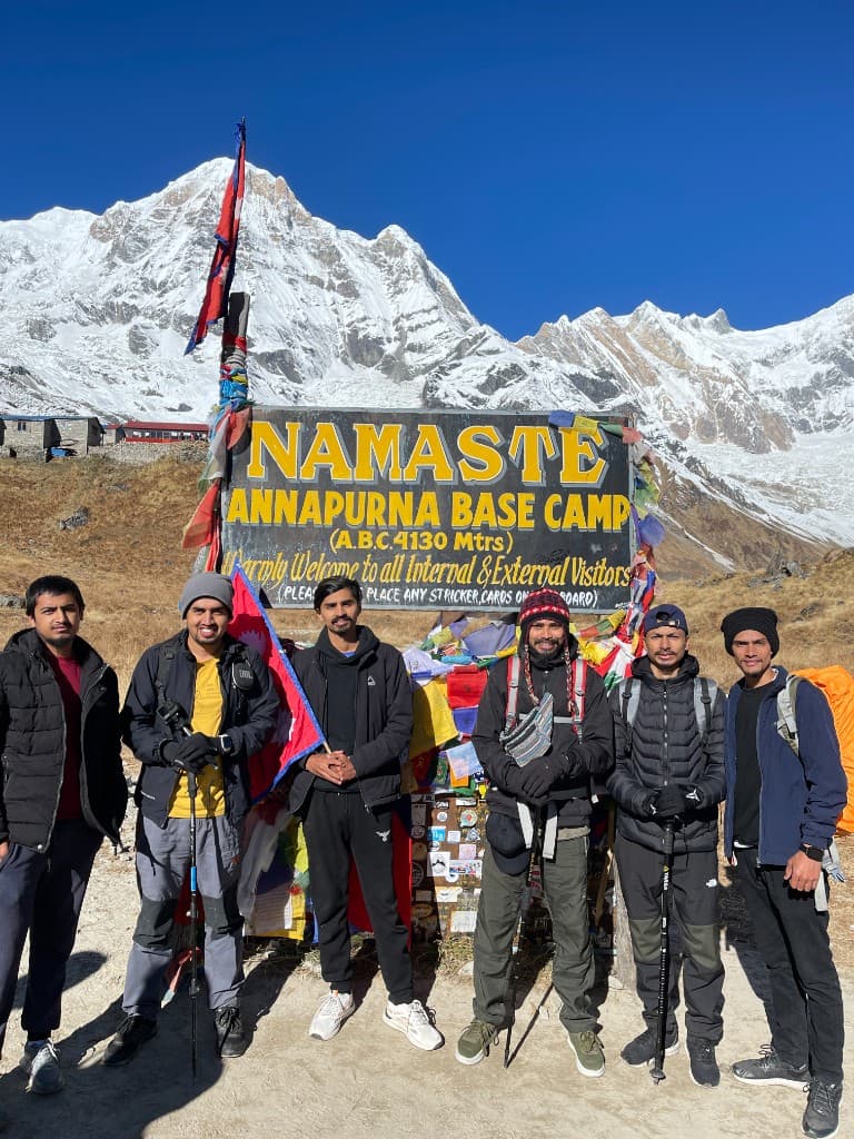 Six friends at the Annapurna Base Camp welcome sign with snow peaks behind