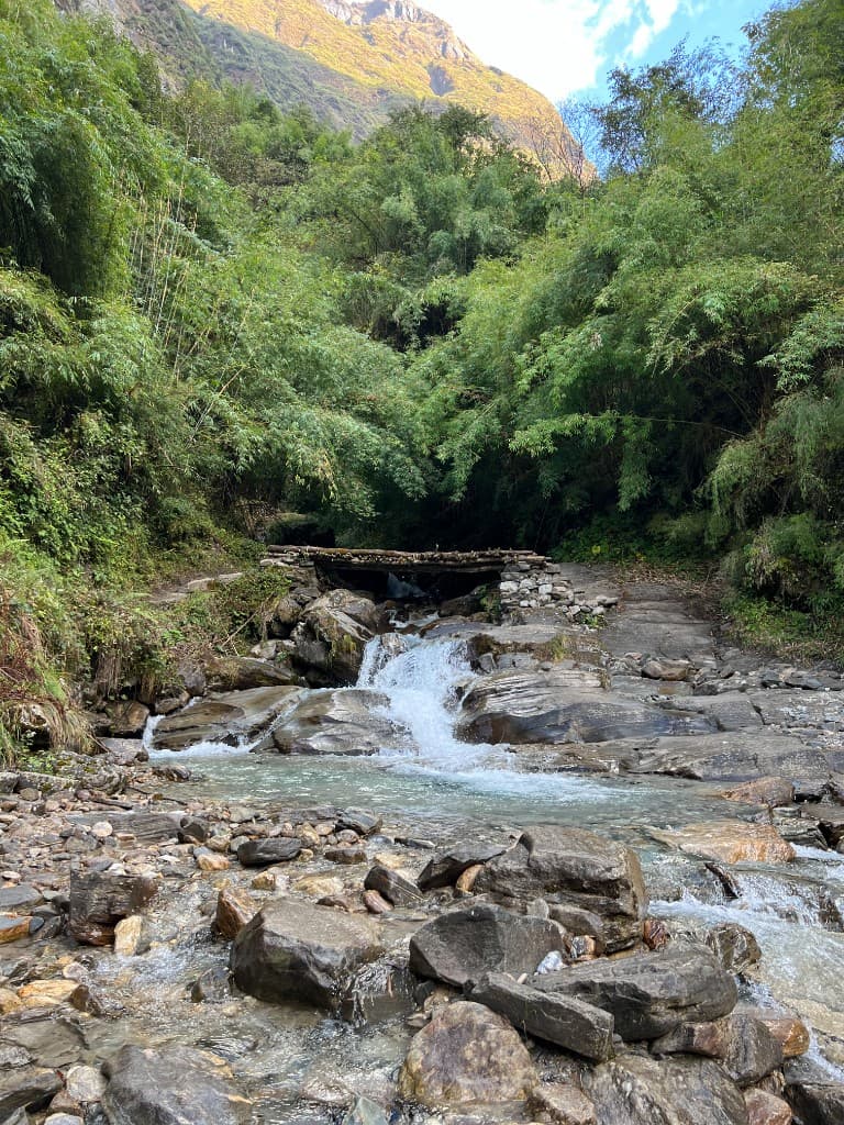 Wooden log bridge crossing a cascading stream with a sunlit peak beyond
